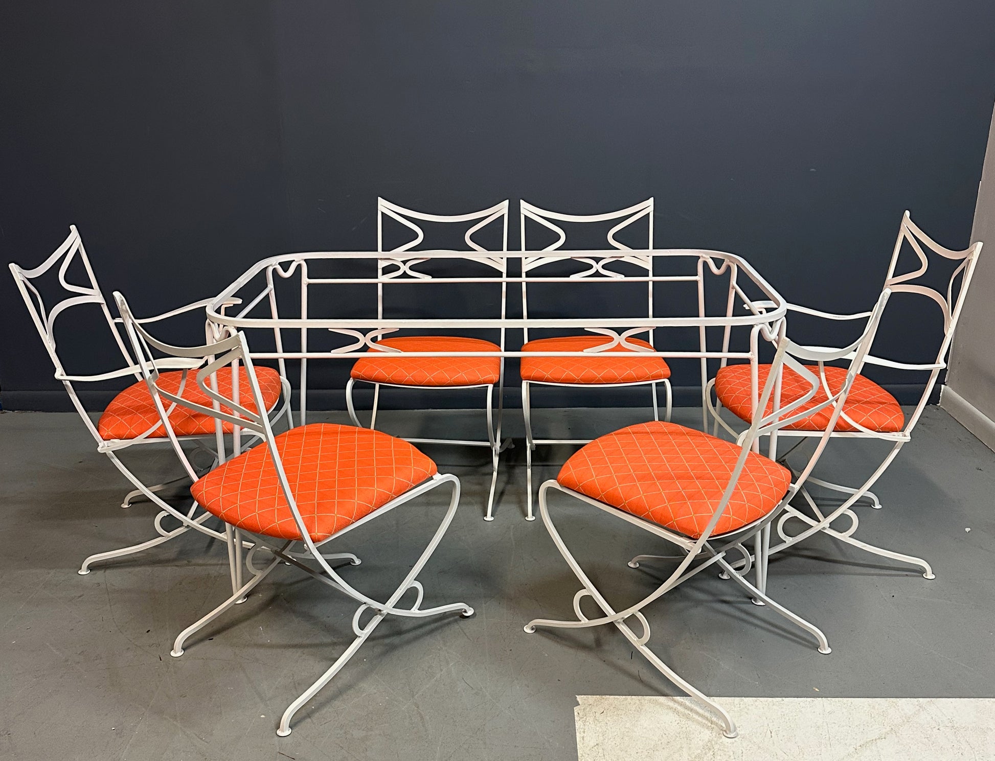 1940s French mid-century modern outdoor dining set featuring six white wrought iron chairs with orange diamond-patterned cushions and a matching table with decorative curved frames