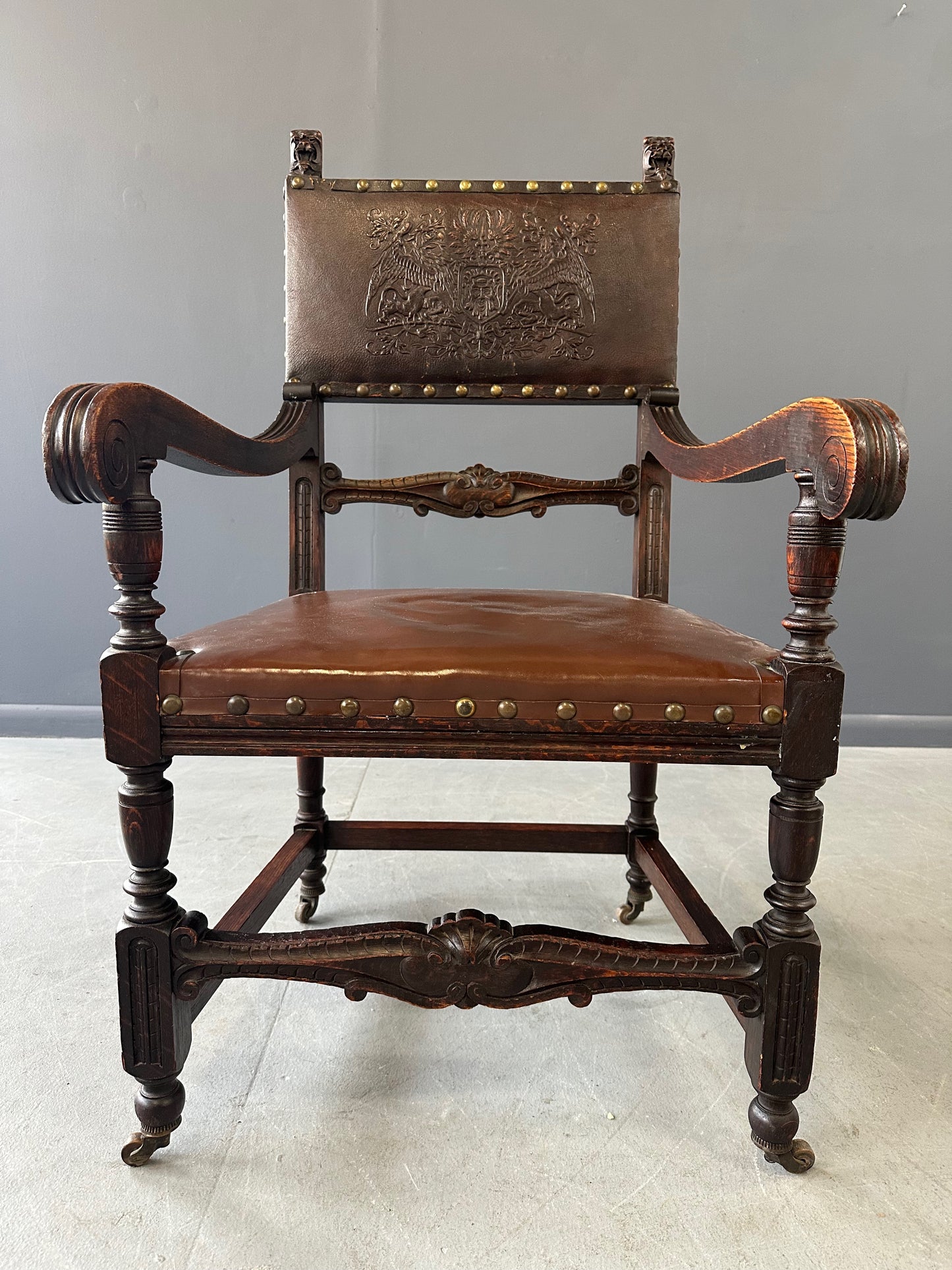 Impressive English 18th-century oak carved armchair featuring tooled leather backrest with heraldic motif, nailhead trim, and ornate turned wood detailing