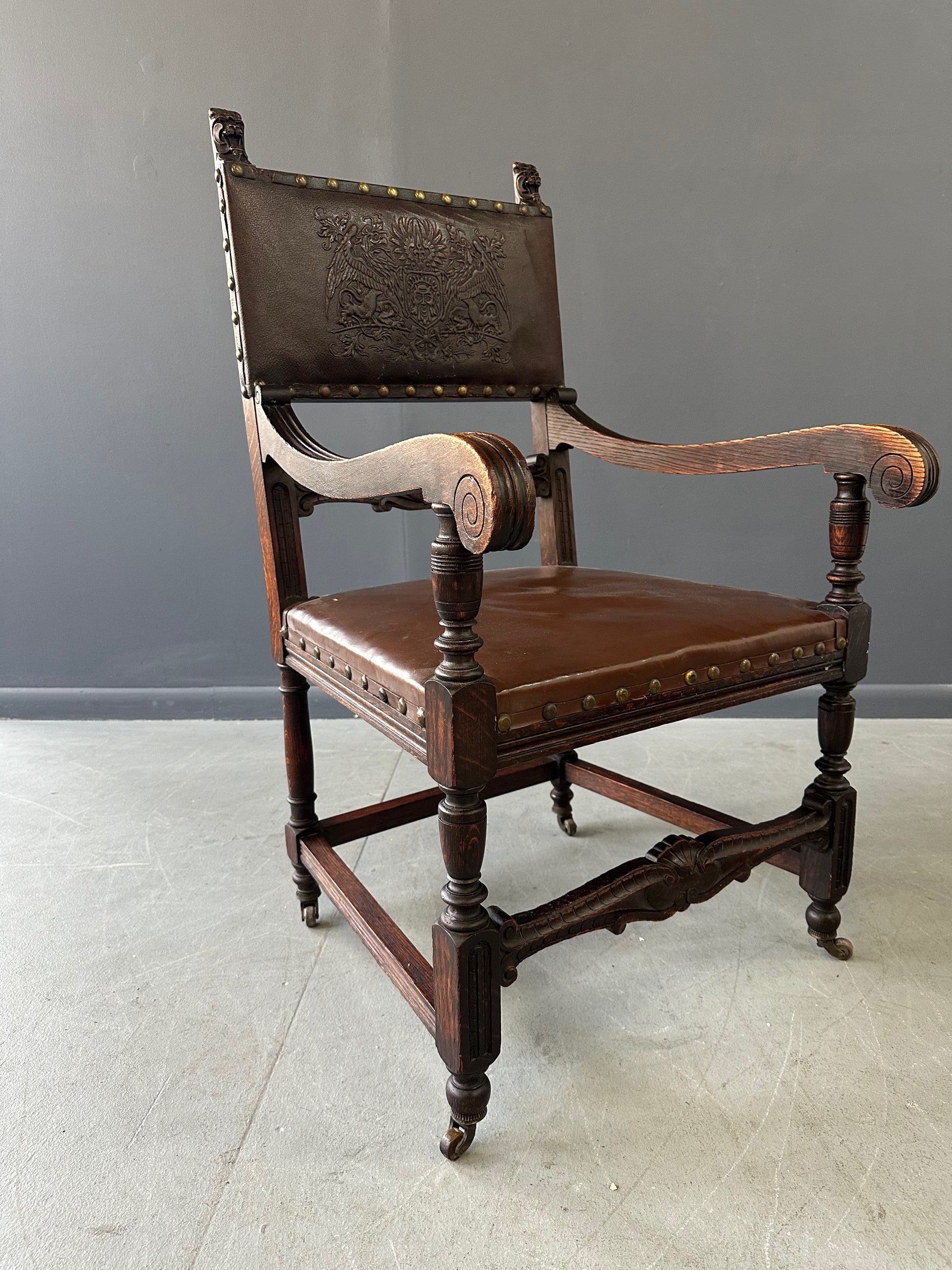 Impressive English 18th-century oak carved armchair featuring tooled leather backrest with heraldic motif, nailhead trim, and ornate turned wood detailing