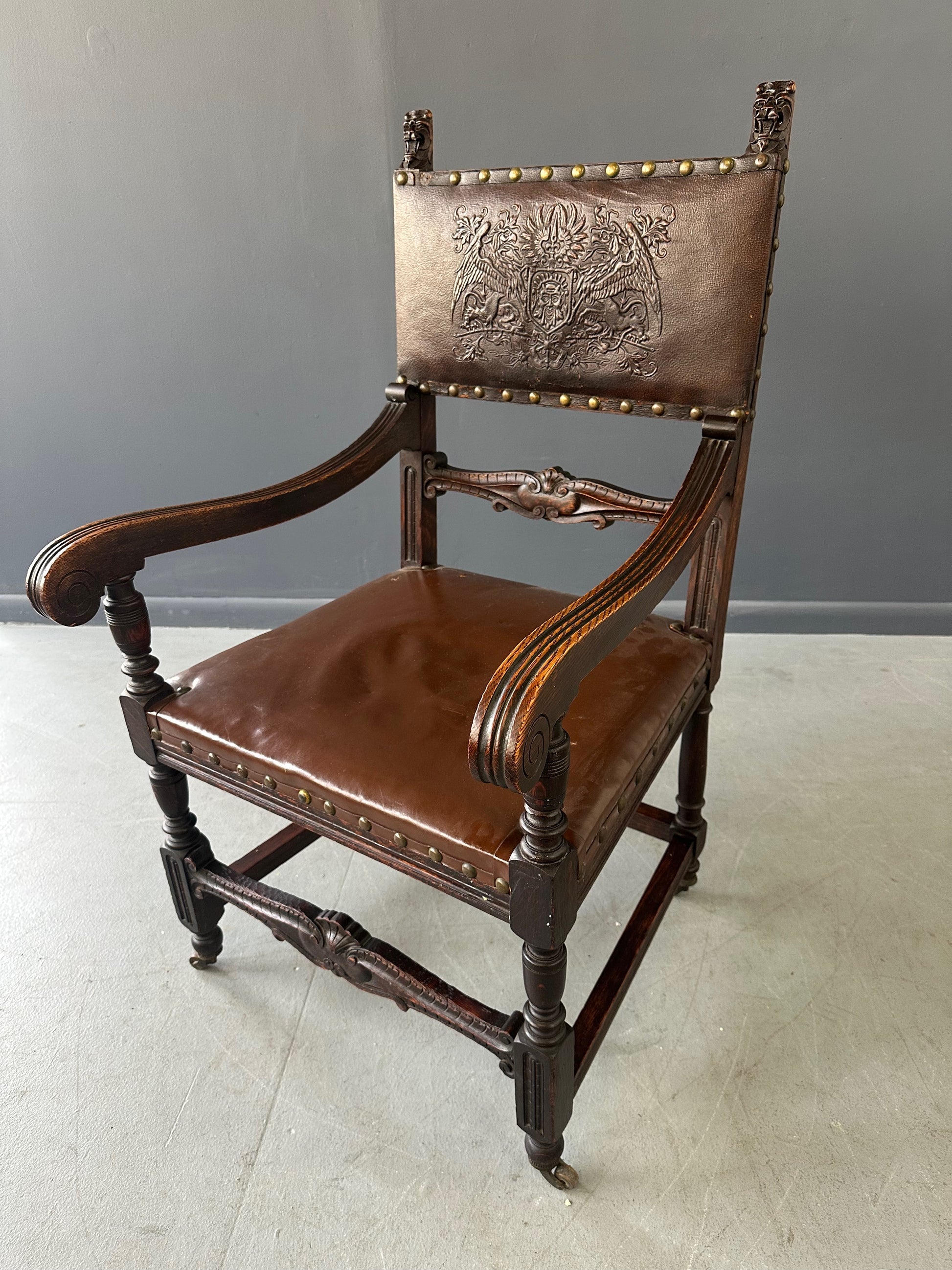 Impressive English 18th-century oak carved armchair featuring tooled leather backrest with heraldic motif, nailhead trim, and ornate turned wood detailing