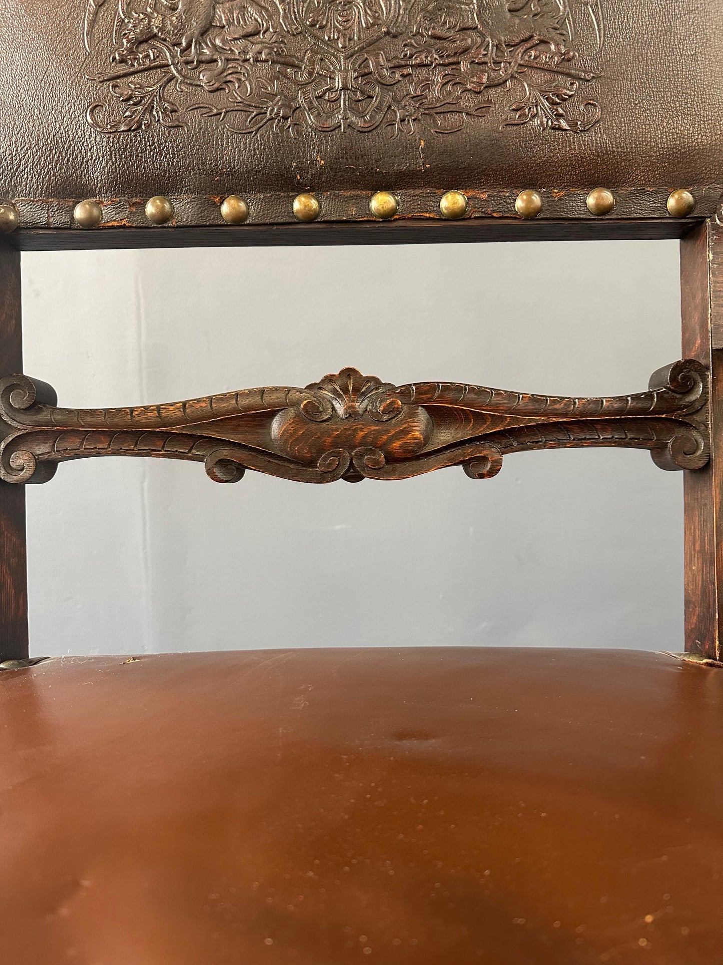 Impressive English 18th-century oak carved armchair featuring tooled leather backrest with heraldic motif, nailhead trim, and ornate turned wood detailing
