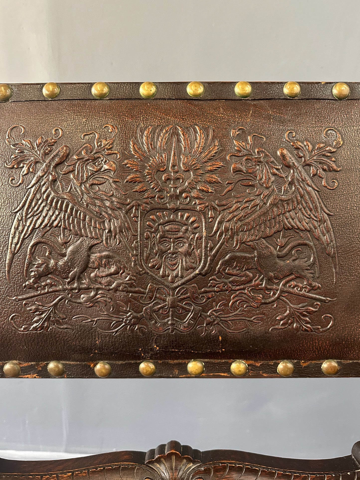 Impressive English 18th-century oak carved armchair featuring tooled leather backrest with heraldic motif, nailhead trim, and ornate turned wood detailing