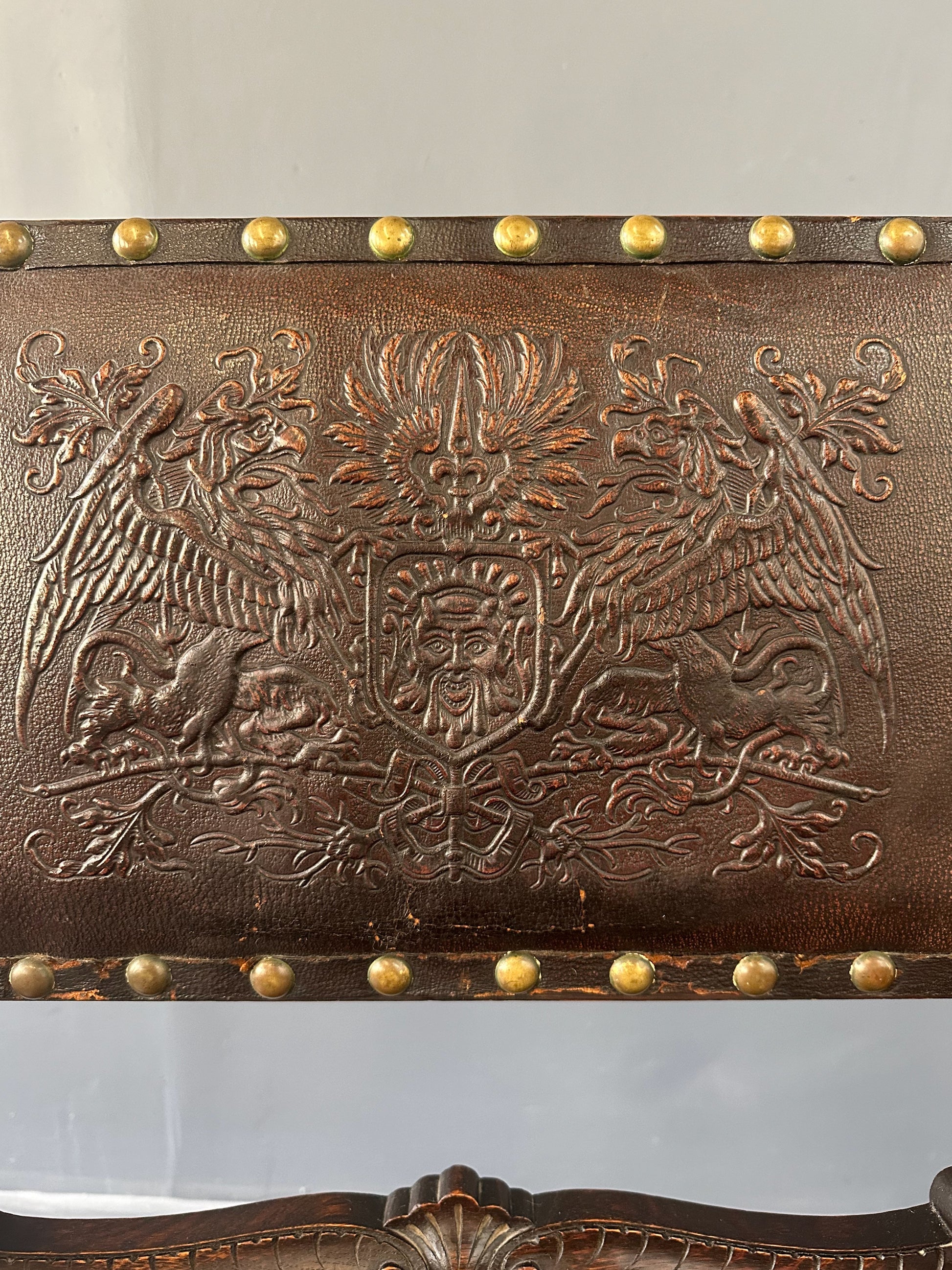 Impressive English 18th-century oak carved armchair featuring tooled leather backrest with heraldic motif, nailhead trim, and ornate turned wood detailing