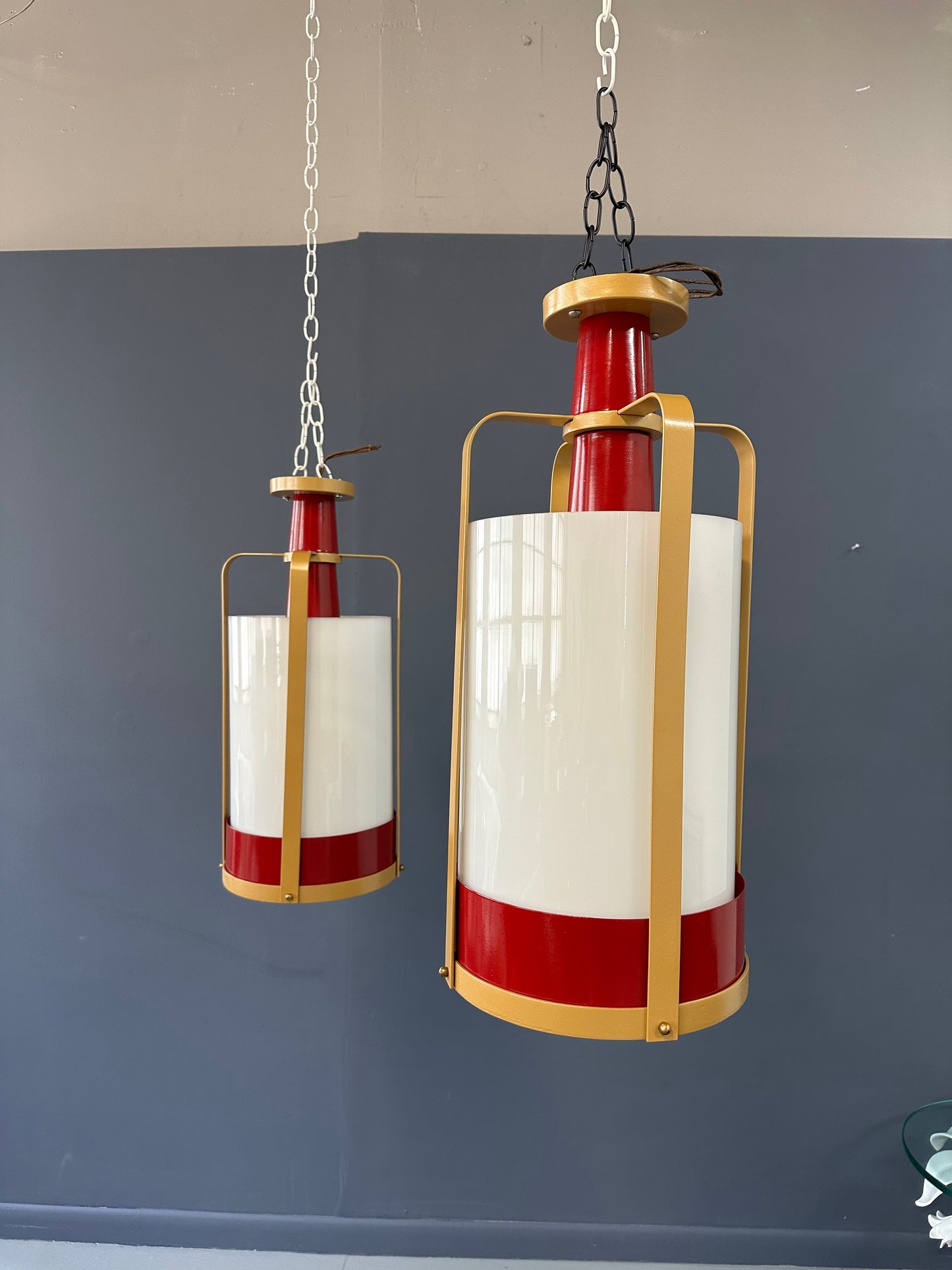 Pair of large pendant lights featuring cylindrical white glass shades encased in red and brass-colored metal frames, suspended by chains for ceiling mounting