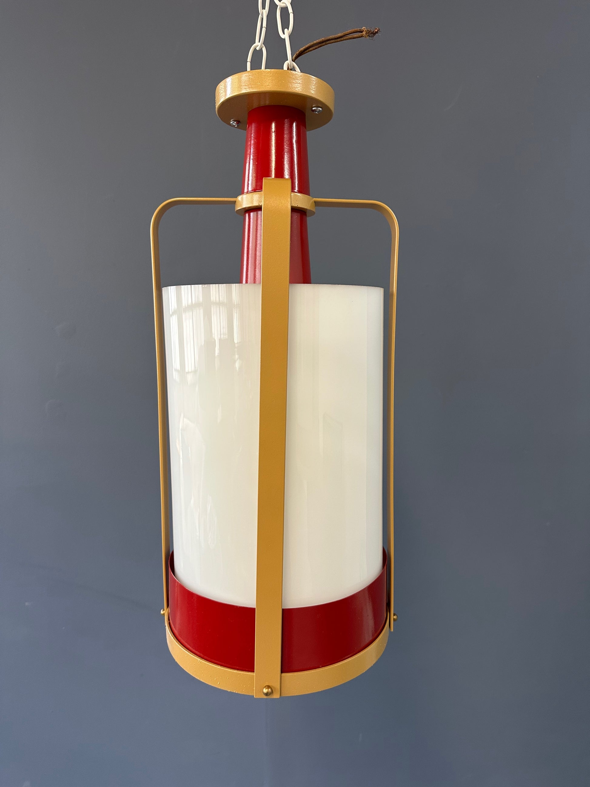 Pair of large pendant lights featuring cylindrical white glass shades encased in red and brass-colored metal frames, suspended by chains for ceiling mounting