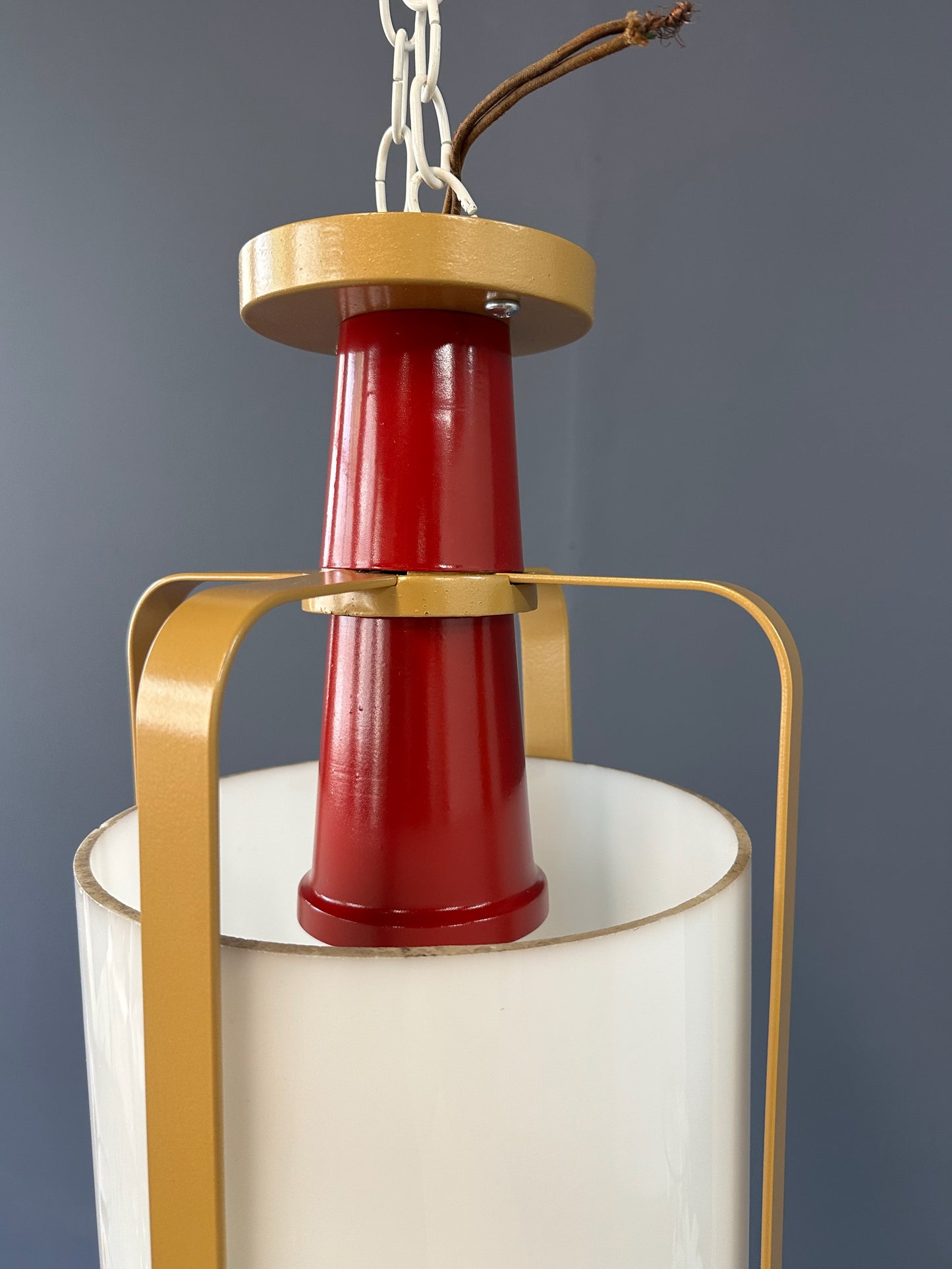 Pair of large pendant lights featuring cylindrical white glass shades encased in red and brass-colored metal frames, suspended by chains for ceiling mounting