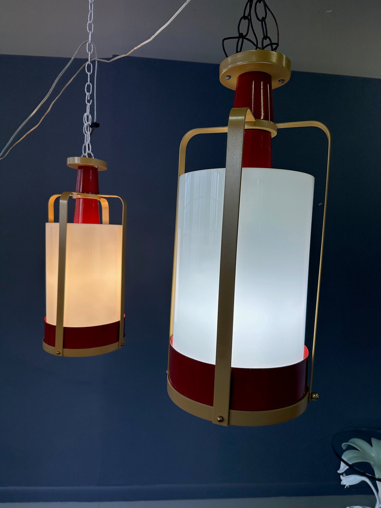 Pair of large pendant lights featuring cylindrical white glass shades encased in red and brass-colored metal frames, suspended by chains for ceiling mounting