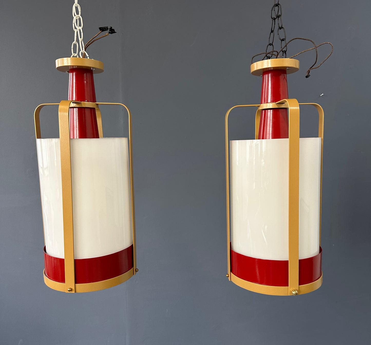 Pair of large pendant lights featuring cylindrical white glass shades encased in red and brass-colored metal frames, suspended by chains for ceiling mounting