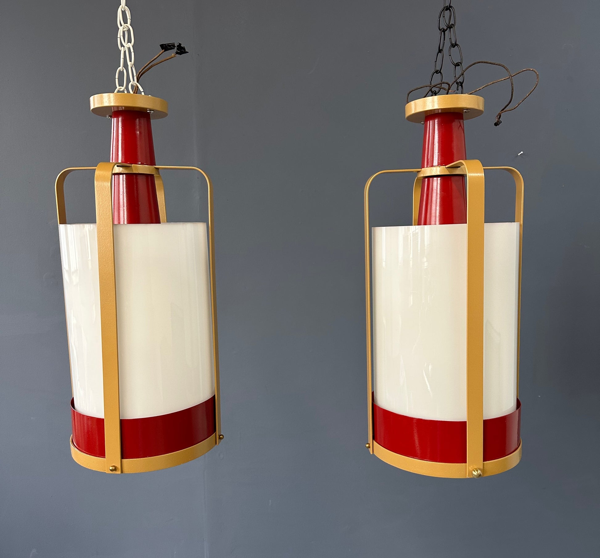 Pair of large pendant lights featuring cylindrical white glass shades encased in red and brass-colored metal frames, suspended by chains for ceiling mounting