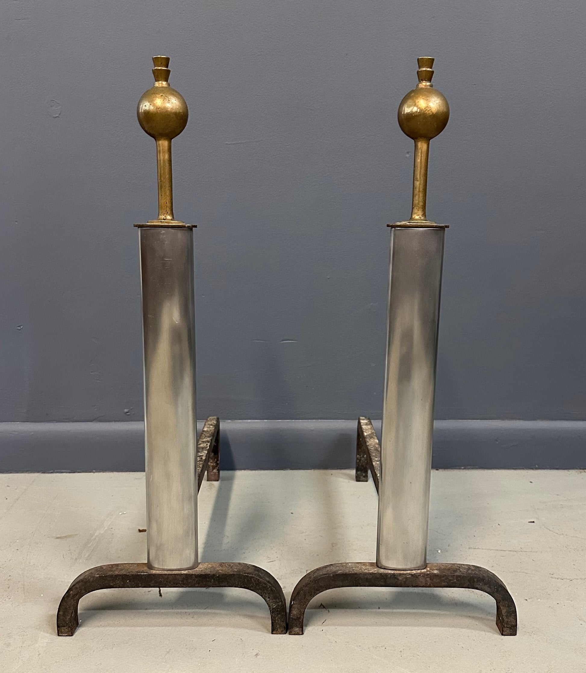 Pair of Machine Age andirons made of polished steel columns with spherical brass finials on top, supported by rugged iron bases with a minimalist, industrial design.