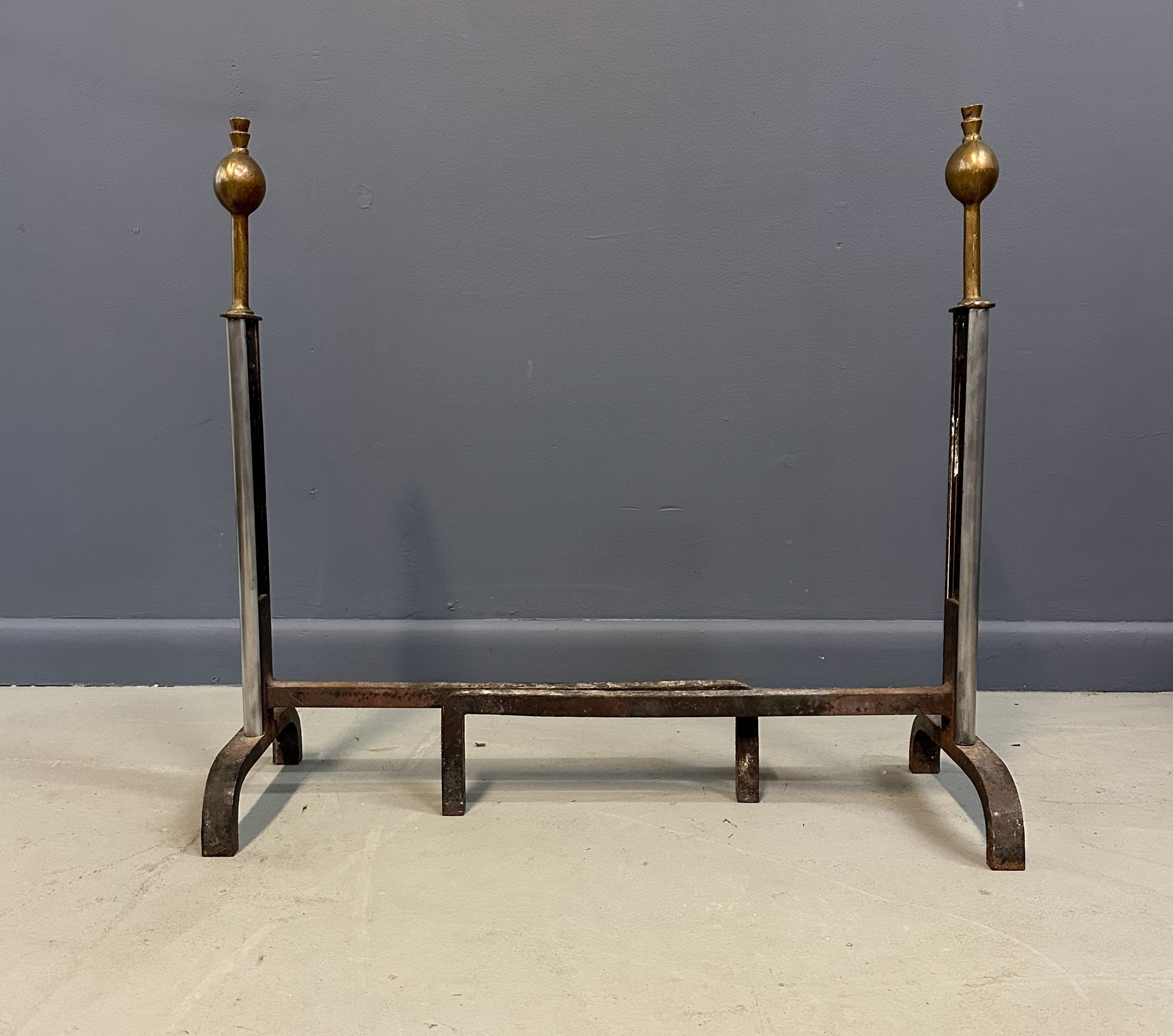 Pair of Machine Age andirons made of polished steel columns with spherical brass finials on top, supported by rugged iron bases with a minimalist, industrial design.
