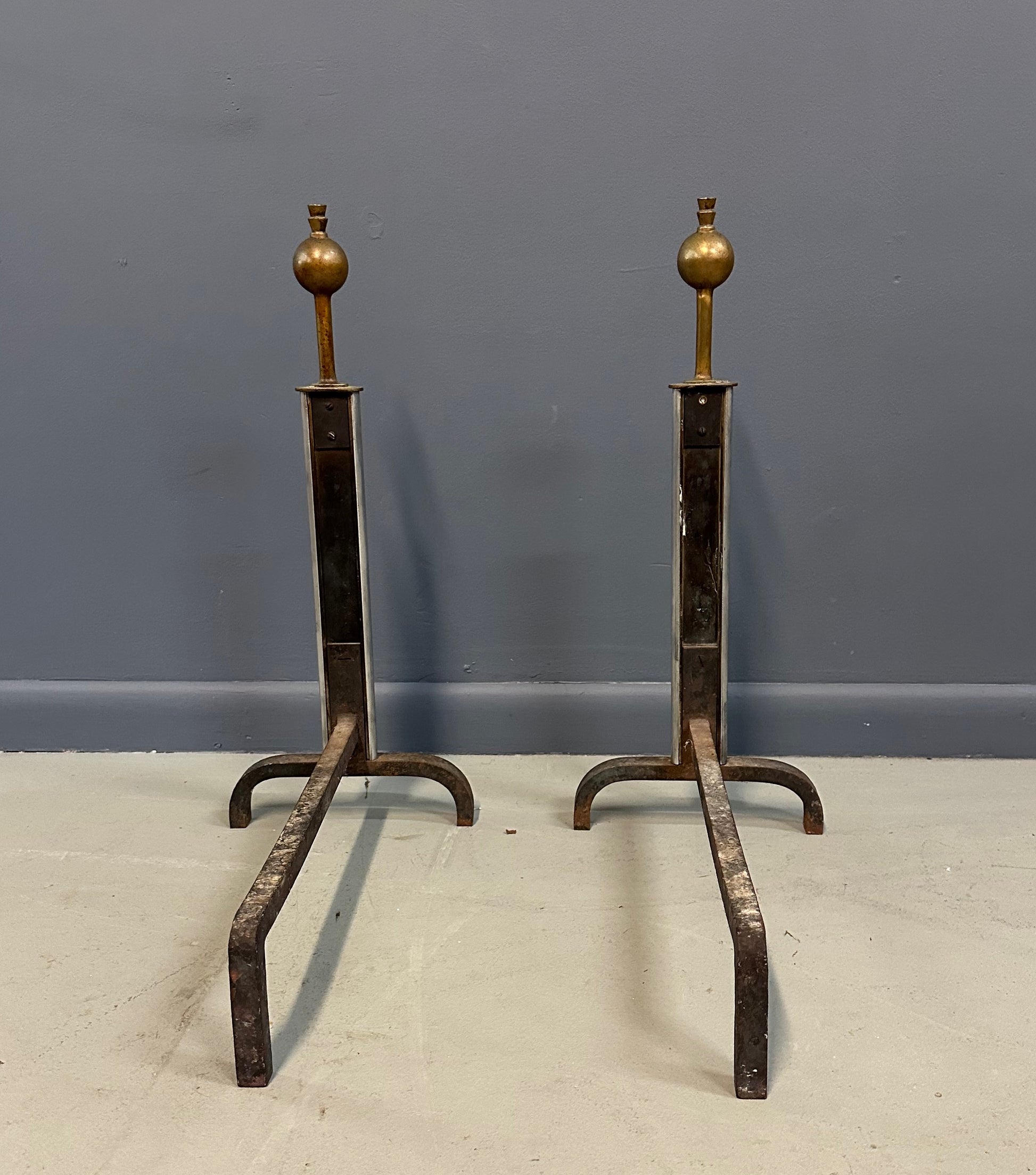 Pair of Machine Age andirons made of polished steel columns with spherical brass finials on top, supported by rugged iron bases with a minimalist, industrial design.