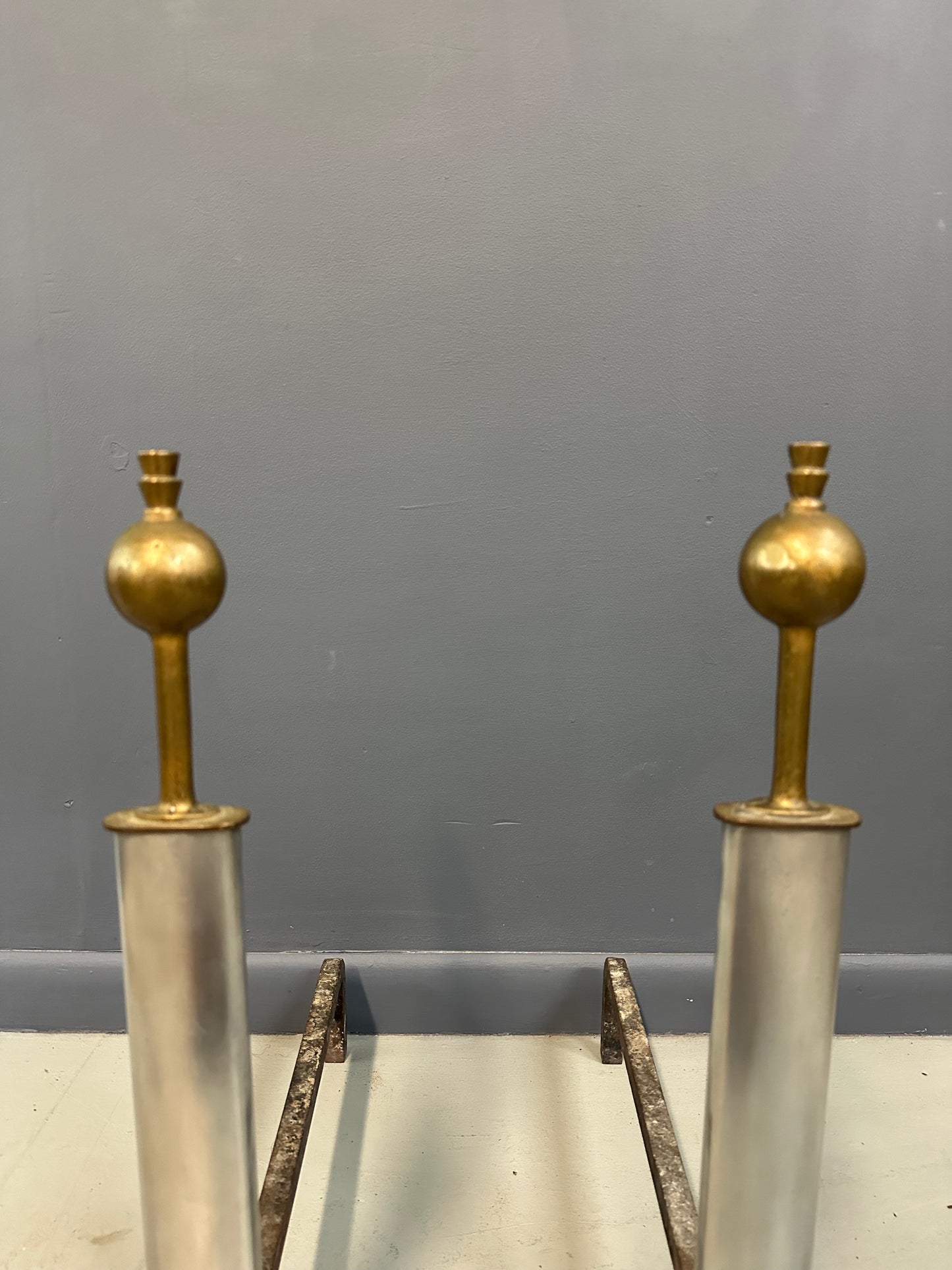 Pair of Machine Age andirons made of polished steel columns with spherical brass finials on top, supported by rugged iron bases with a minimalist, industrial design.